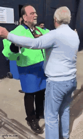 A hi-vis-vested venue steward performing an exaggerated, theatrical pat-down of a man walking past, all show and no actual search.